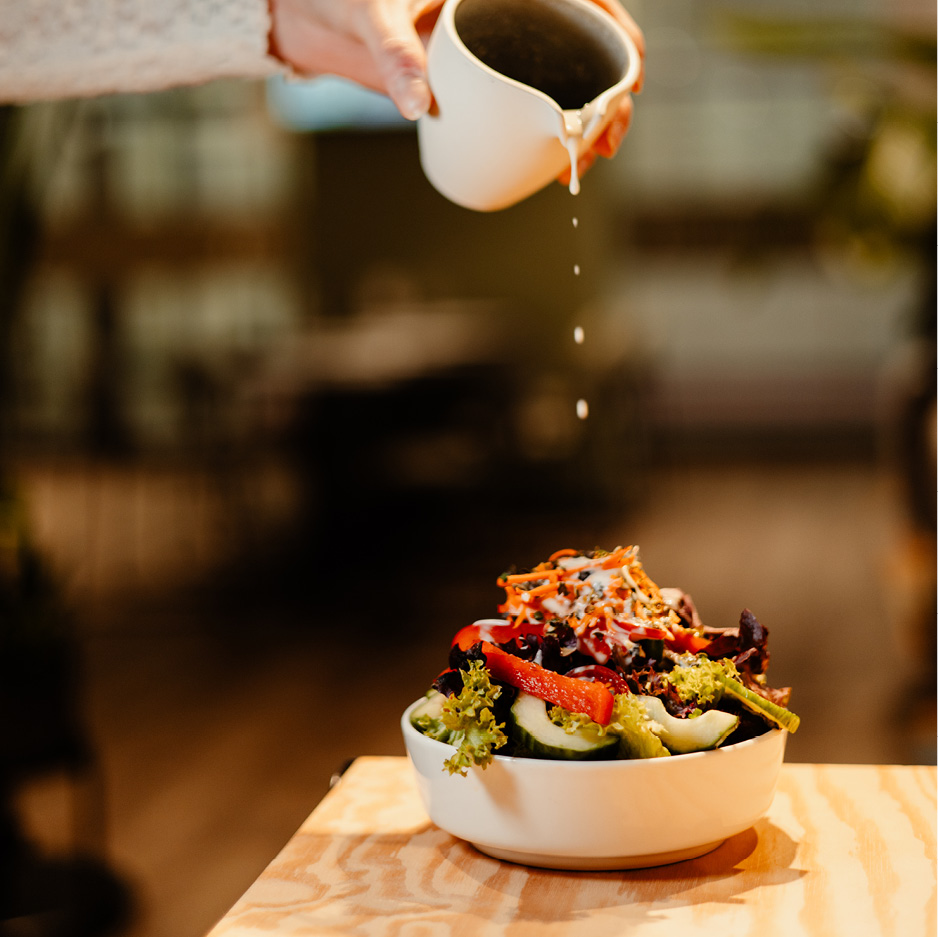 Das Bild zeigt einen bunt gemischten Beilagensalat, über den jemand ästhetisch Dressing gießt.
