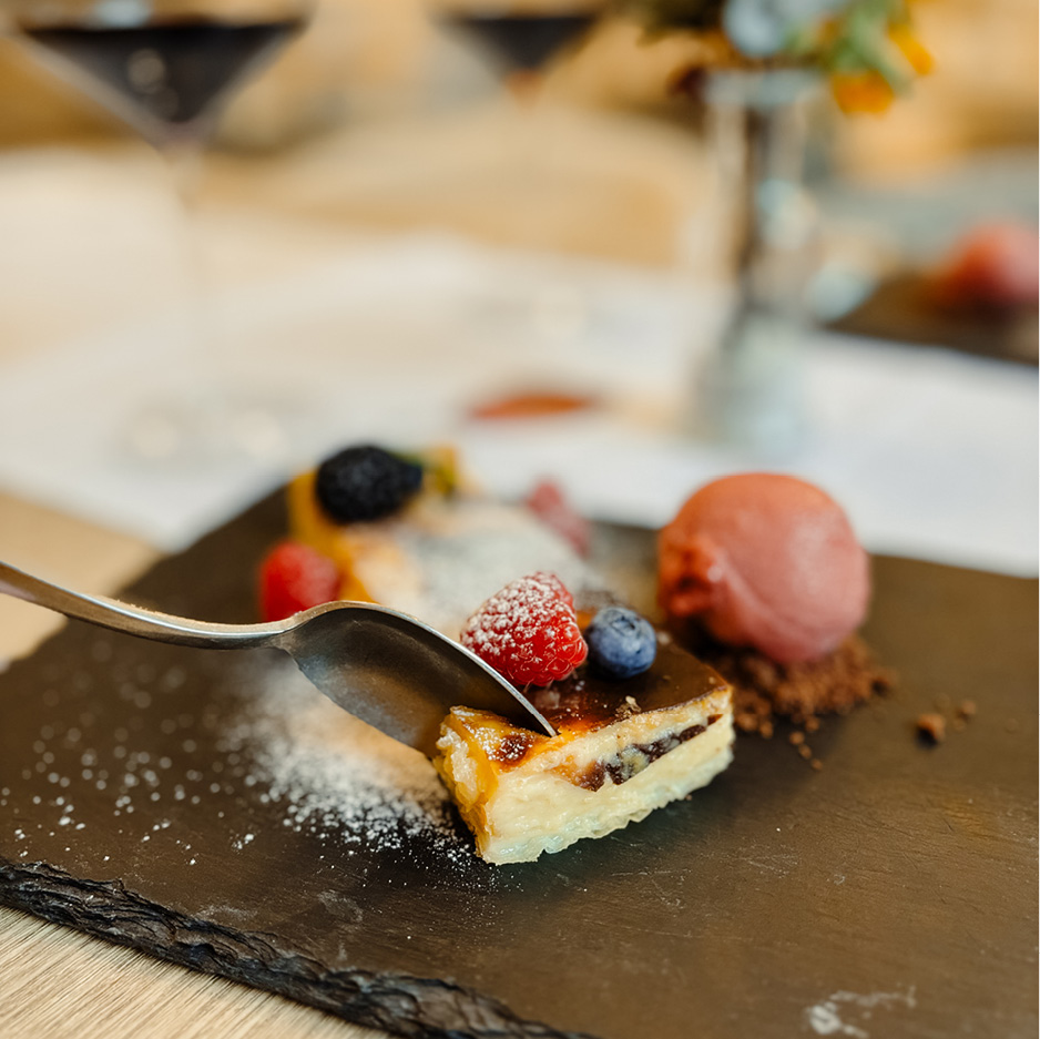 Das Bild zeigt ein köstliches Dessert auf einer dunklen Schieferplatte, in das gerade ein Löffel sticht.