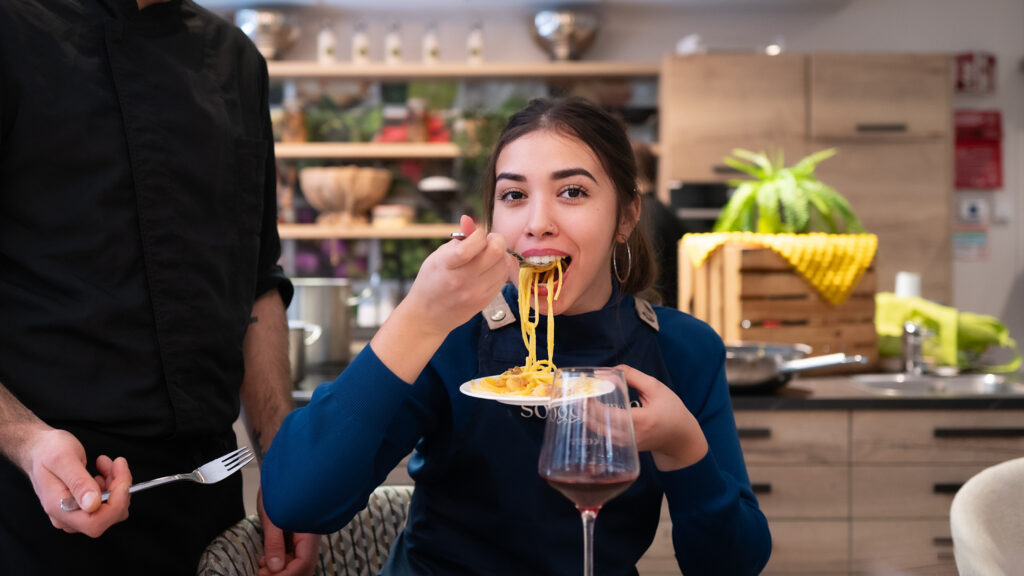 Das Bild zeigt eine junge Frau in der kulinario-Kochschule, die begeistert Pasta löffelt.