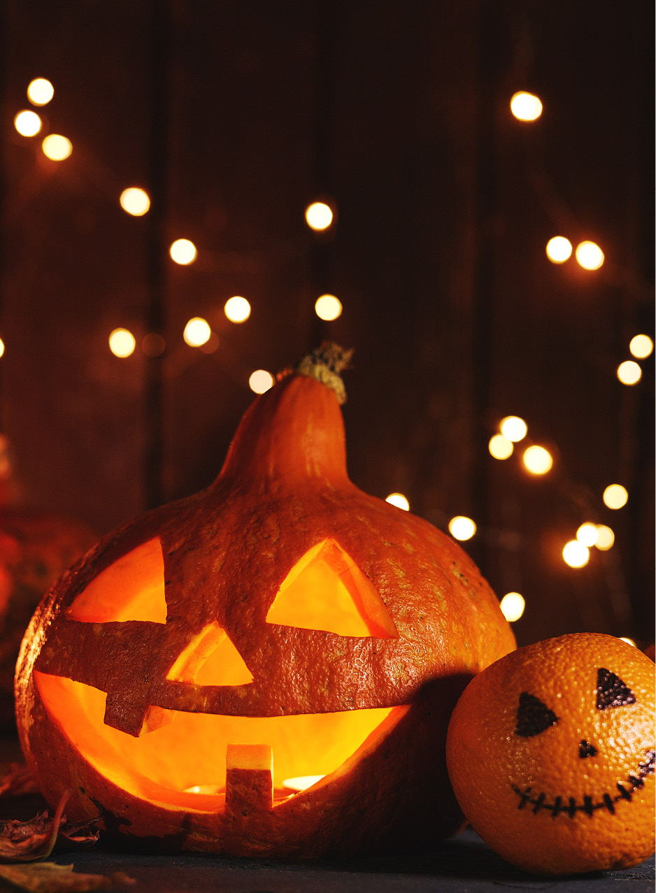 Das Bild zeigt einen geschnitzten Halloweenkürbis und Mandarinen mit Gesichtern vor dunklem Glühwürmchen-Hintergrund.