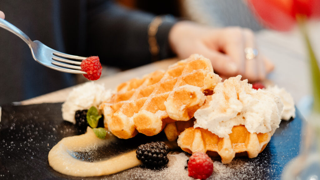 Das Bild zeigt eine Zimt-und-Zucker-Waffel auf einer dunklen Schieferplatte und eine von der Gabel aufgespießte Himbeere.