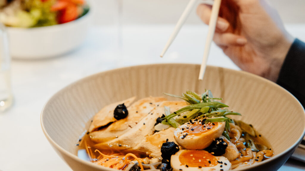 Das Bild zeigt das Hendl-Ramen-Gericht von kulinario, das eine Frau gerade mit Stäbchen verspeist.