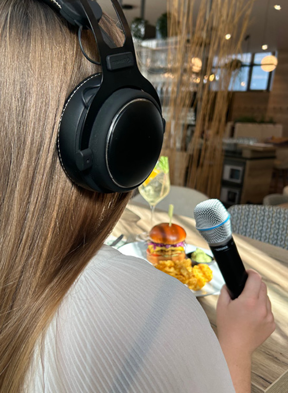 Das Bild zeigt eine Frau von schräg hinten mit Kopfhörern und Mikrofon bei PodKost, vor ihr ein Burger.